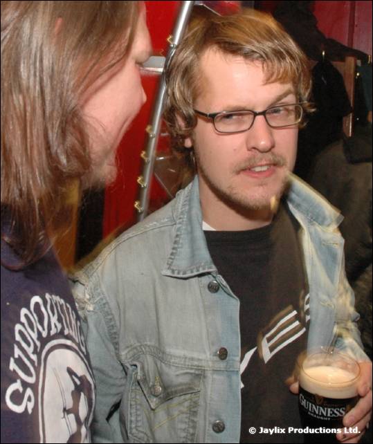 A man with glasses and a denim jacket holds a pint of Guinness, with another person in the foreground