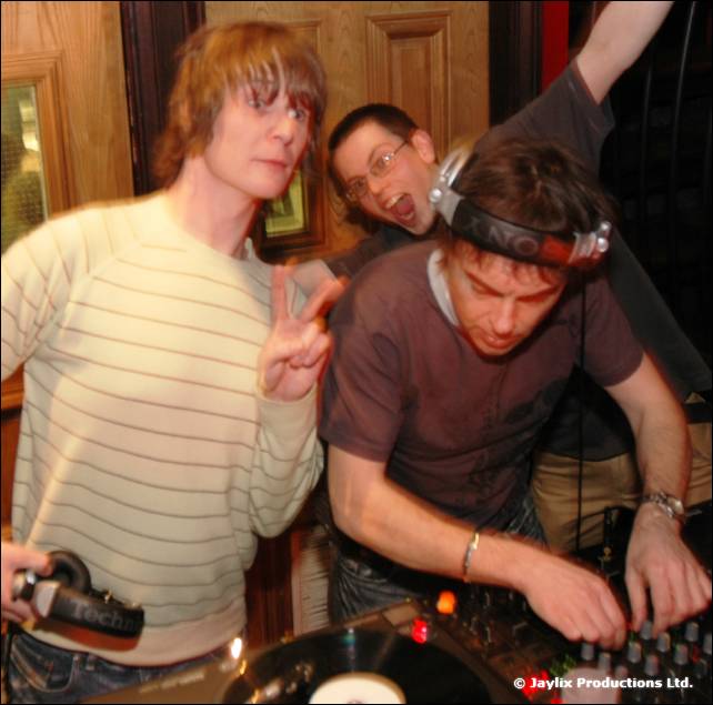 Two people at DJ decks, one in headphones and one making a peace sign, with a third person behind