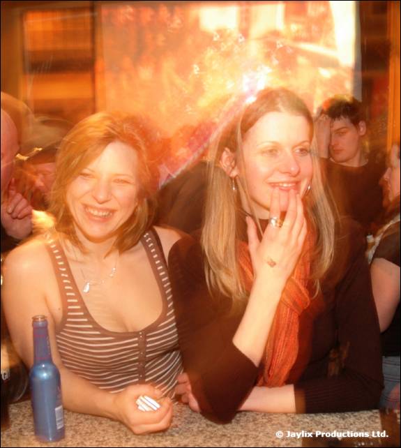 Two women lean on a bar smiling, with a blue bottle in front of them and a glowing orange background