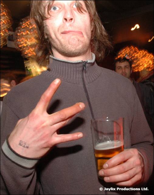 A man in a grey zip top pulls a face and holds up three fingers, with a pint of lager in the other hand
