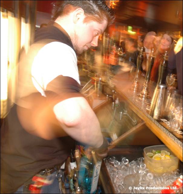 A bartender leans over the bar preparing drinks, with a tub of ice and lemon slices below