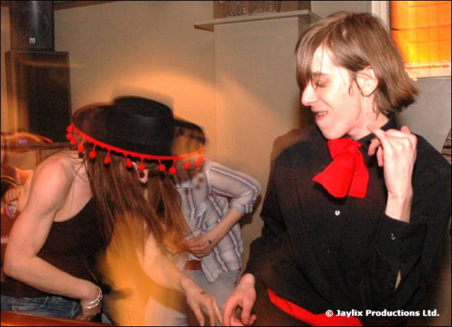 A person in a wide black hat with red pom poms and a person in a black shirt with a red bow tie dance