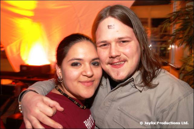A woman in a dark red top and a man with long hair and a mark on his forehead smile together