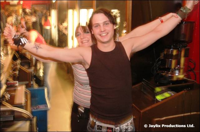 A smiling man in a dark vest top holds his arms wide at a bar, with a woman behind him