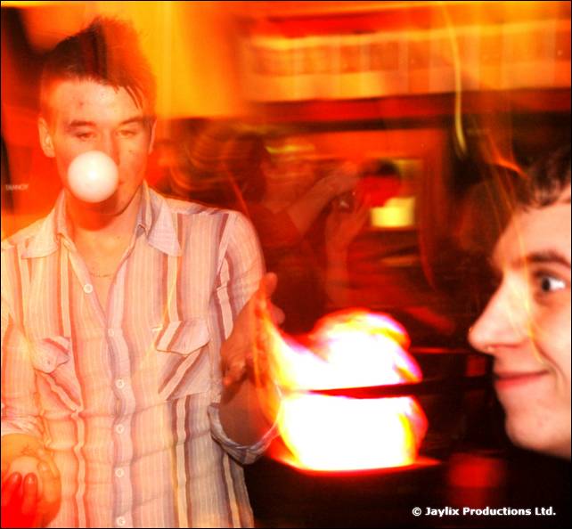 A man in a striped shirt balances a white ball on his nose above a flaming dish, with another face at the edge