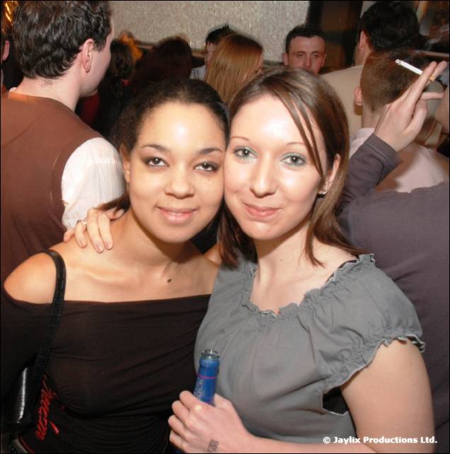Two women pose together in a crowded bar, one holding a blue bottle