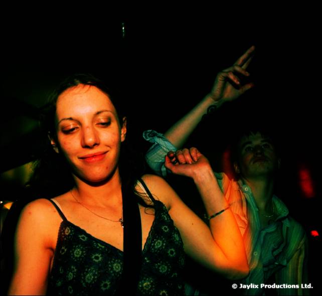 A woman in a dark floral top dances with one arm raised in a dark room, with a person behind her