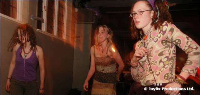 Three women dance in a room with large windows, one wearing glasses and a patterned jacket