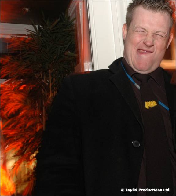 A man in a dark suit and tie screws up his face in front of a plant, lit in orange
