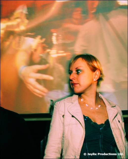 A woman in a cream jacket looks away to the side, in front of a blurred projected image