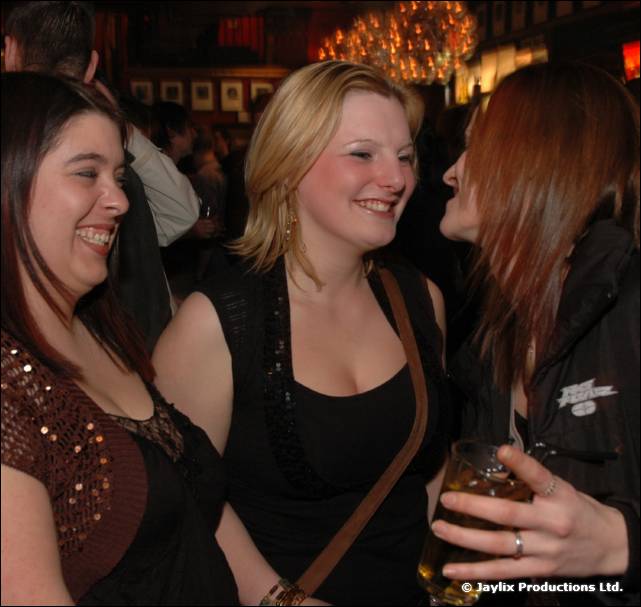Three women laugh and talk close together in a busy bar, one holding a drink