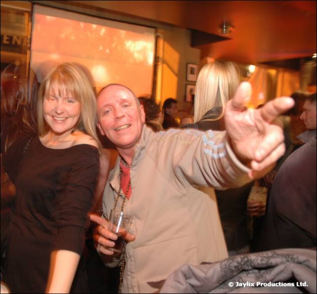 A woman and a man in a beige jacket pose in a bar, the man pointing towards the camera