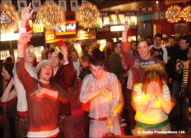 A crowd with arms raised in a bar, with chandeliers above and a woman covering her face in the foreground