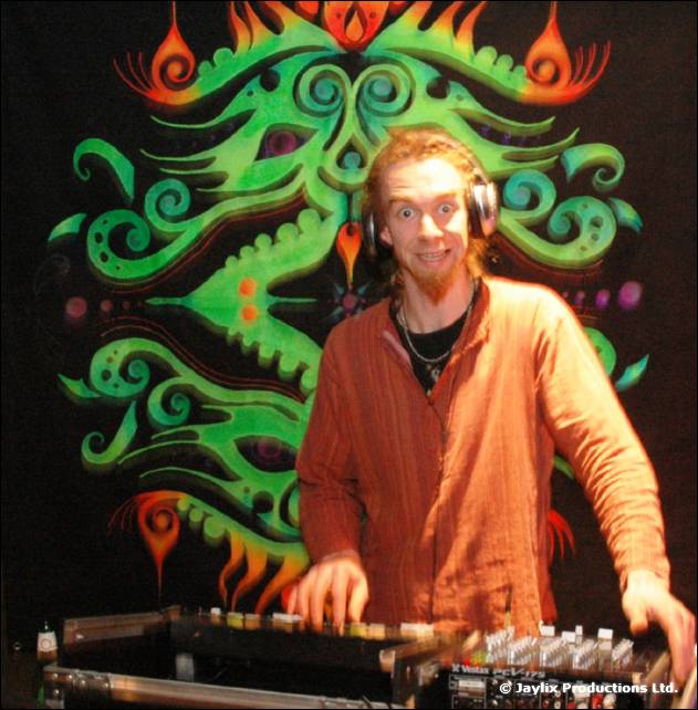 A DJ in headphones grins at the camera at decks, in front of a green and orange tribal backdrop