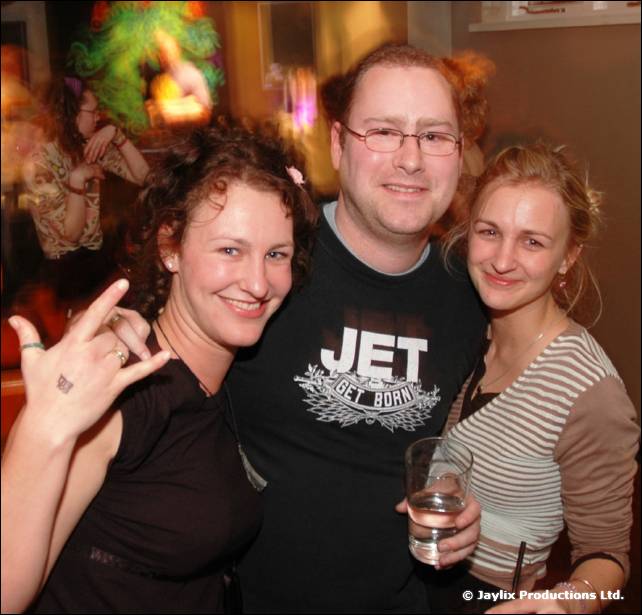 A man in glasses and a Jet t-shirt poses with two women in a bar, one holding up a hand