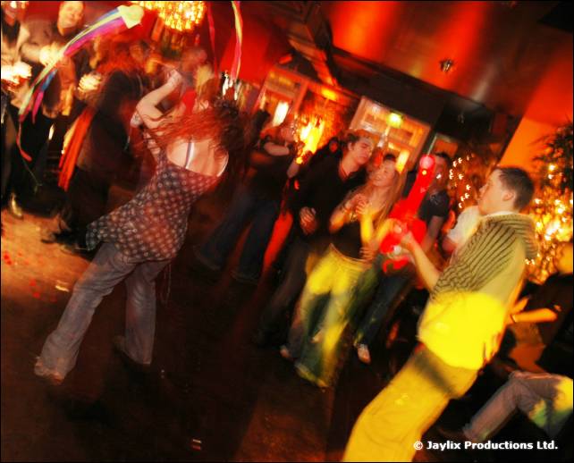 People dance in a blurred, warmly lit room, with coloured ribbons swinging at the top