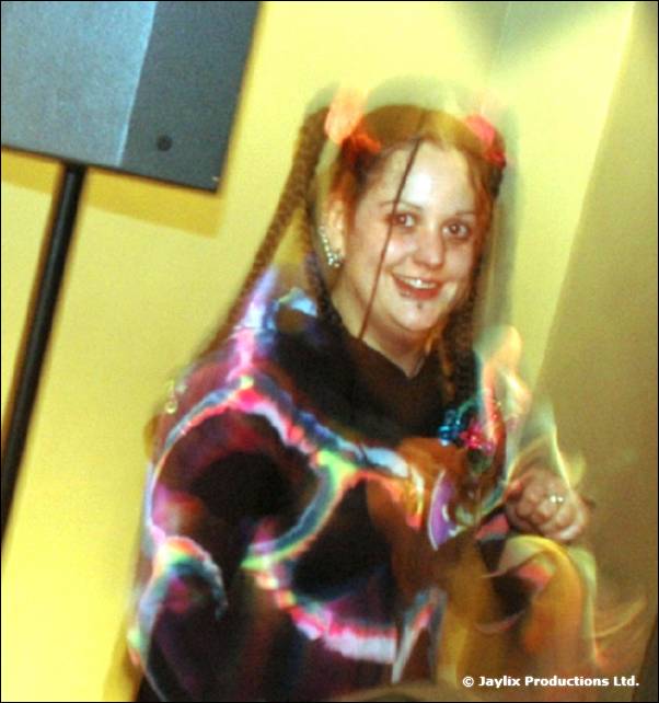 A person with bunches smiles, sitting in front of a yellow wall, with a glowing pattern of light trails on their top