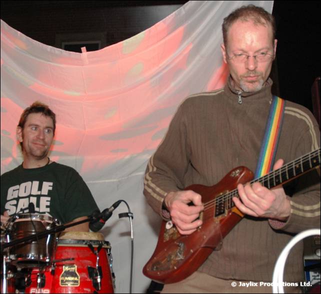 A man plays a red bass guitar with a rainbow strap beside a drummer in a Golf Sale t-shirt