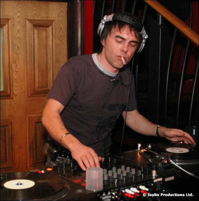 A DJ in headphones with a cigarette in his mouth mixes at decks in front of a wooden door