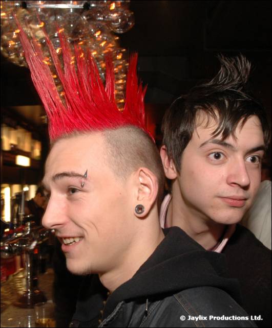 A person with a red mohican and a piercing in the eyebrow, beside a person with dark spiky hair