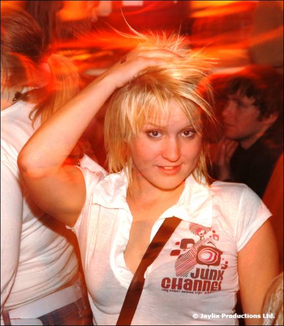 A woman with short blonde hair holds one hand to her head, wearing a white Junk Channel t-shirt