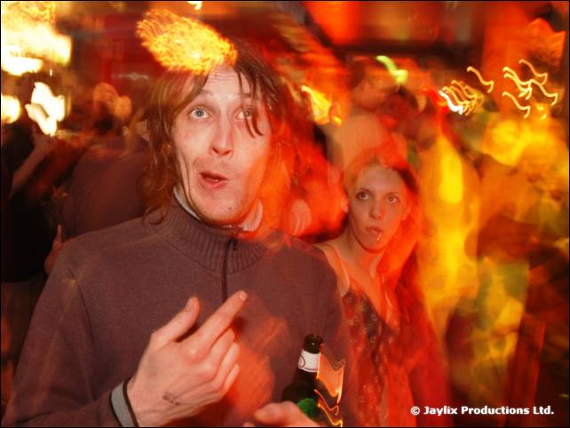 A man in a grey zip top pulls a face and points, in a busy bar with blurred orange lights