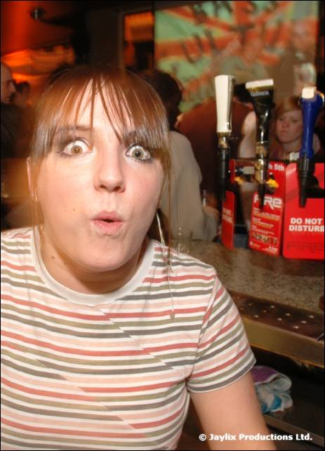 A woman in a striped t-shirt leans on a bar with wide eyes, beside beer taps and a FIRE flyer