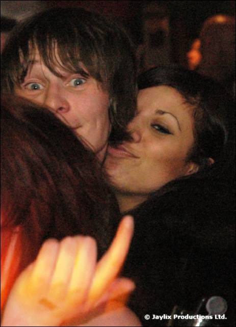 Two people embrace cheek to cheek, one pulling a face, with a hand held up in the foreground