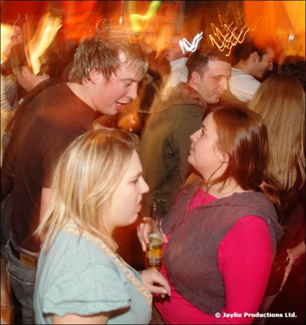 Two women and a man talk in a crowded bar, with light streaks above