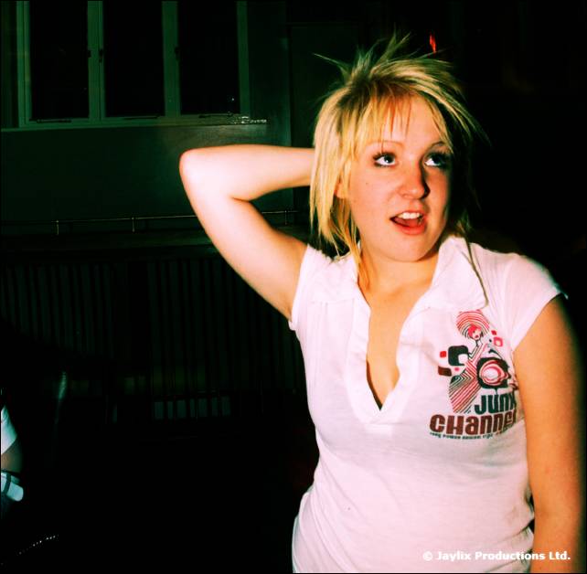 A woman with short blonde hair poses with one hand behind her head, wearing a Junk Channel t-shirt, in a dark room