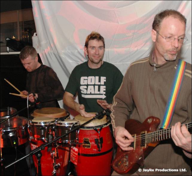 A band plays on a stage, with a drummer, a man in a Golf Sale t-shirt on congas and a guitarist