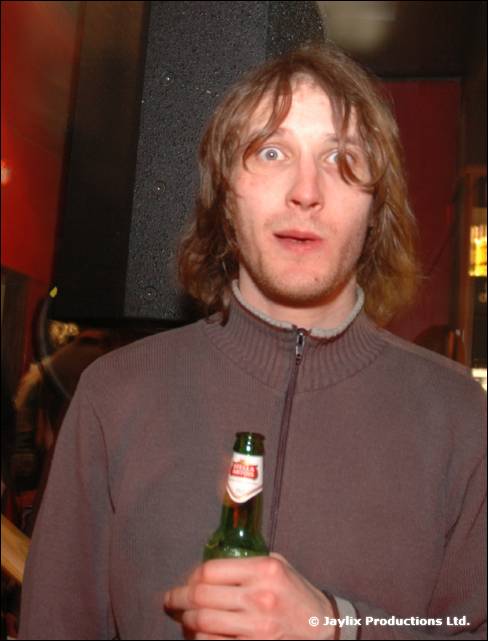 A man with long hair and a grey zip top holds a green beer bottle, looking wide eyed at the camera