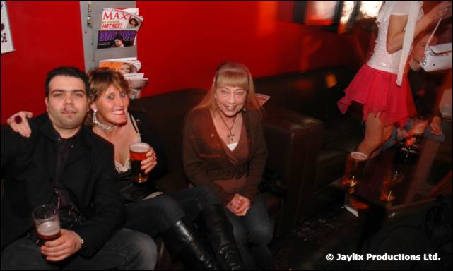 A man and two women sit on a black sofa with drinks, against a red wall