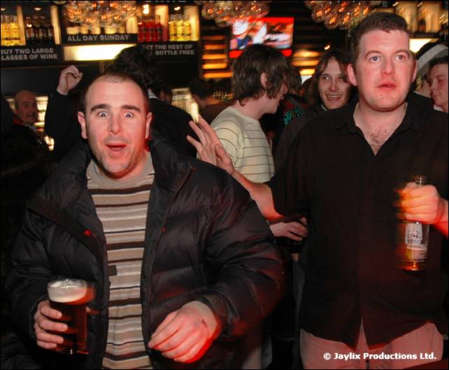 Two men in a bar, one in a puffer jacket holding a pint, the other in a dark shirt holding a bottle