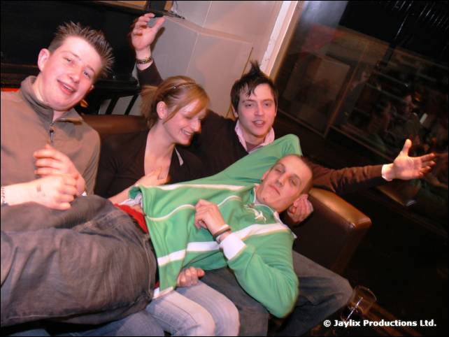 Three people sit on a leather sofa while a man in a green top lies across their laps with his arms spread