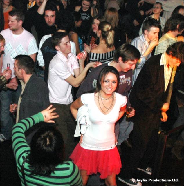 A smiling woman in a white top and red skirt stands among a crowd on a dance floor, seen from above