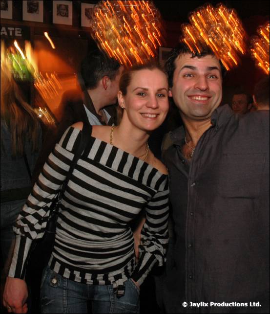 A woman in a striped top and a man in a grey shirt pose together, smiling, in a bar with glowing lights