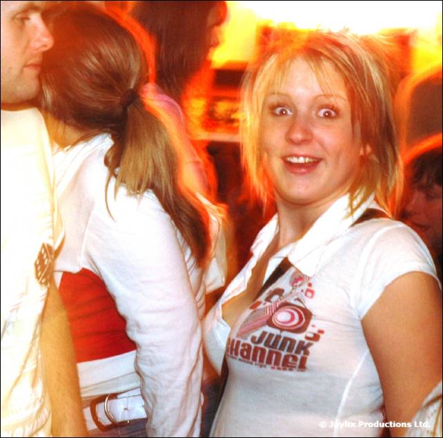 A woman with short blonde hair and wide eyes looks at the camera in a white top, with people behind her