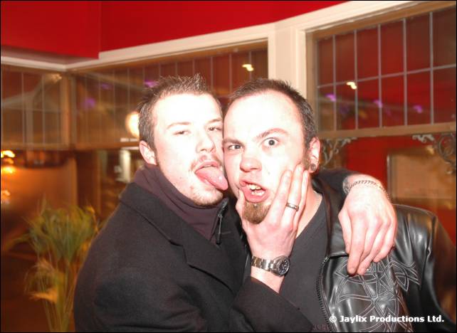 Two men pose cheek to cheek, one sticking his tongue out and the other pulling a face, in a bar