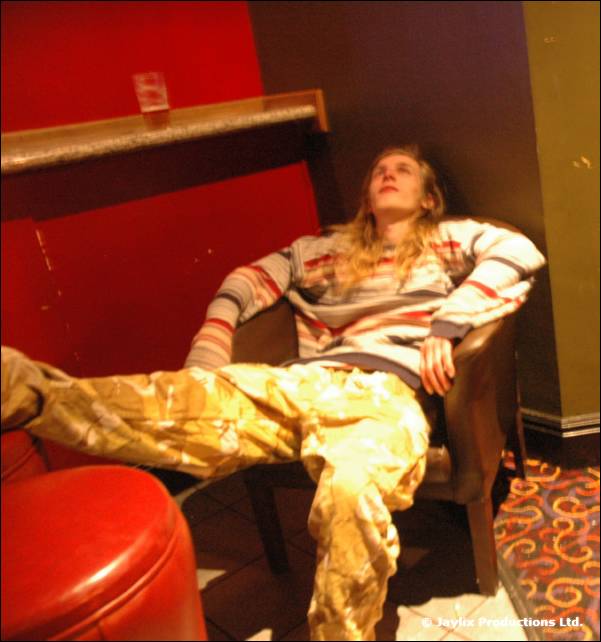 A person with long hair in a striped jumper and camouflage trousers reclines in an armchair beside a red pouffe