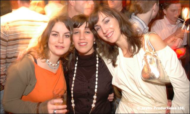 Three smiling women pose close together in a busy bar, one holding a drink