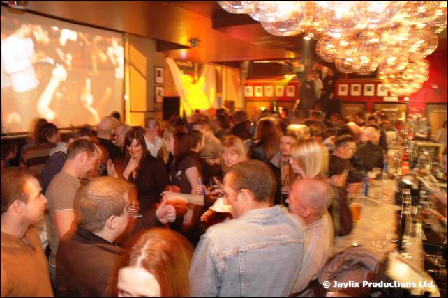A crowded bar with people standing at the counter, a large projection screen and globe lights overhead