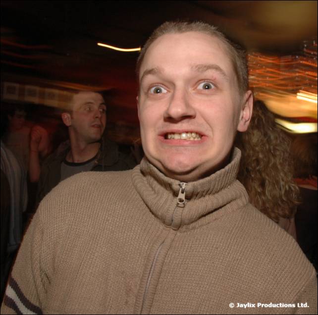 A person in a beige zip-up top pulls a wide-eyed grimace at the camera in a dark club