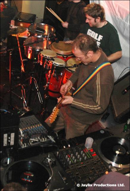 A guitarist with a rainbow strap and a drummer play beside DJ decks and a mixer