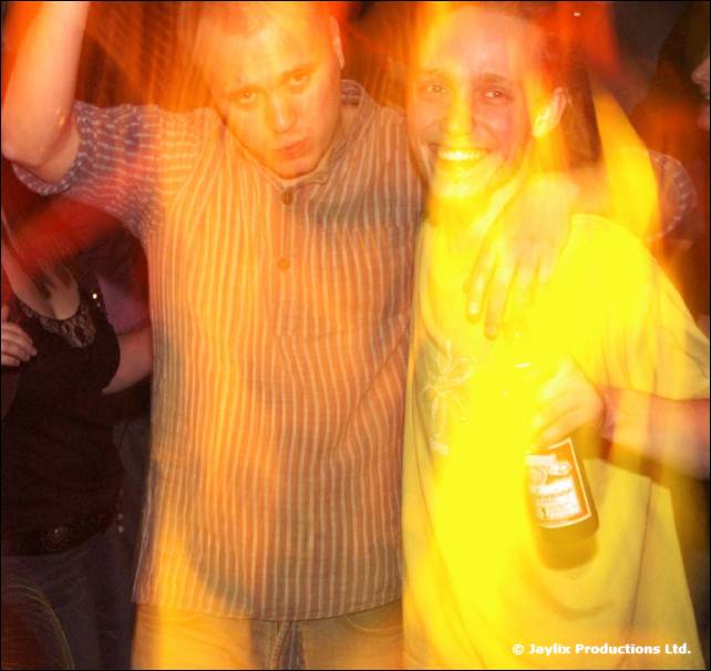 Two men with arms around each other, one in a striped shirt, one in a yellow t-shirt holding a bottle, in blurred warm light