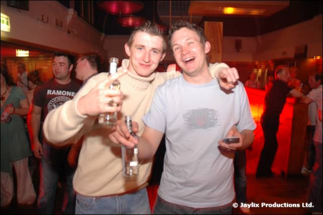 Two men in casual tops hold bottles and smile in a busy venue