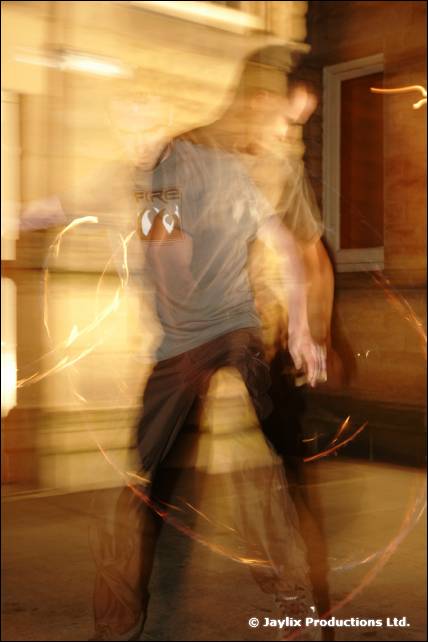 A blurred person in a grey top stands in front of a stone wall with faint light trails