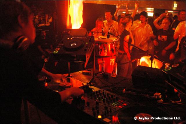 A DJ at the decks in the foreground with a flame effect and drums behind, crowd beyond