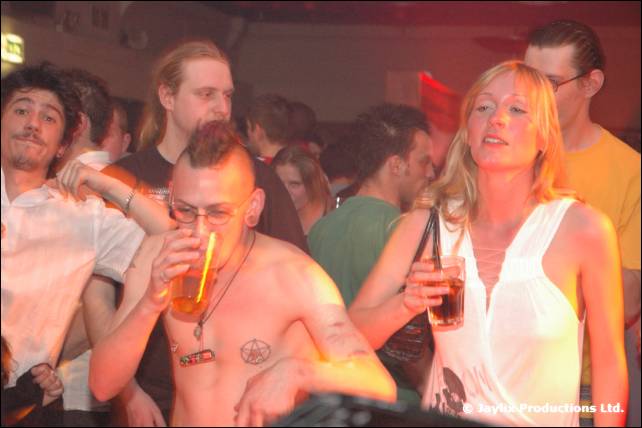 A shirtless man with a mohican drinks in a crowded club, a woman in a white top beside him