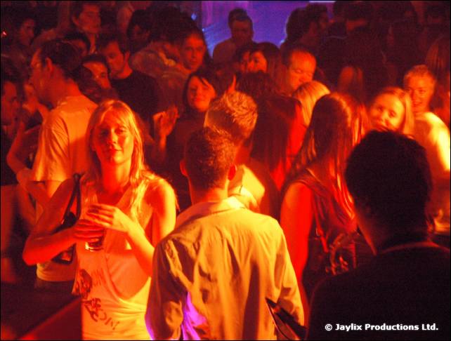 A crowd dances in orange and red light, a blonde woman holding a drink at the front